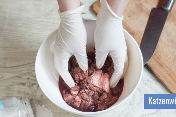 Zwei Hände in Einmalhandschuhen mischen Fleisch in einer Schüssel, daneben liegen ein Schneidebrett aus Holz und ein großes Messer