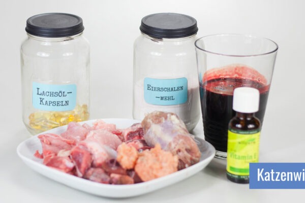 Verschiedene Supplemente für die Rohfütterung der Katze: Muskelfleisch, Innereien und fleischige Knochen auf einem Teller, daneben ein Glas Blut und zwei Schraubgläser mit Bierhefe und Lachsölkapseln