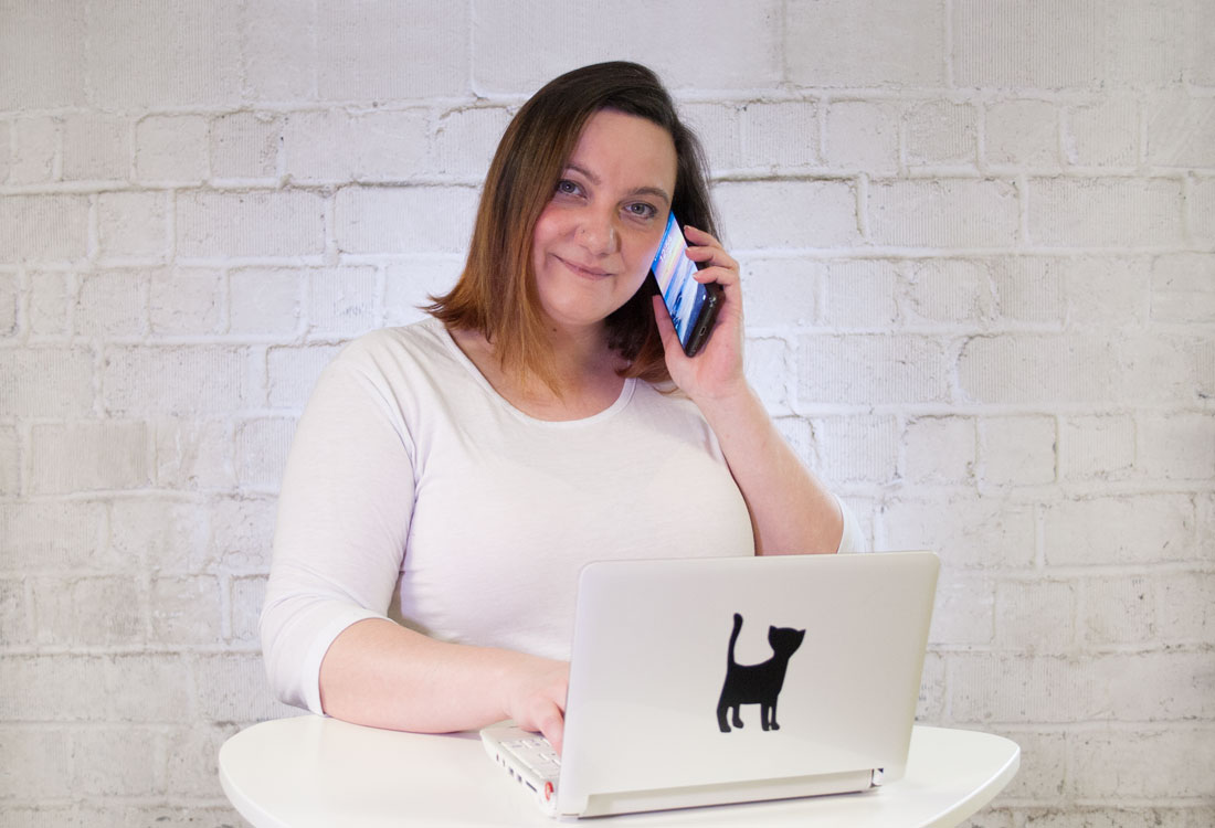 Eine brünette Frau im weißen T-Shirt mit Handy am Ohr steht an einem Stehtisch mit Laptop