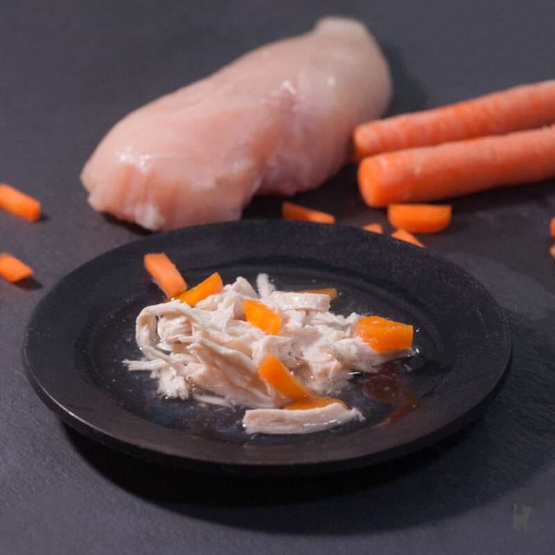 Das Foto zeigt einen Teller mit Schonkost für die Katze. Im Hintergrund liegen eine rohe Hähnchenbrust und Karotten