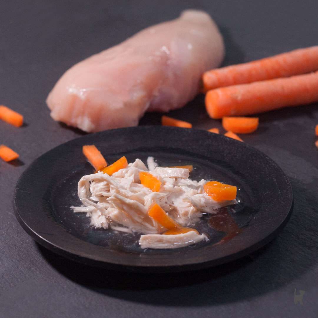 Das Foto zeigt einen Teller mit Schonkost für die Katze. Im Hintergrund liegen eine rohe Hähnchenbrust und Karotten