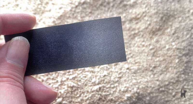 Das Foto zeigt eine Hand, die einen Streifen schwarzen Kartons hält. Der Streifen ist zur Hälfte mit hellem Staub bedeckt. Im Hintergrund ist die Katzenstreu zu sehen