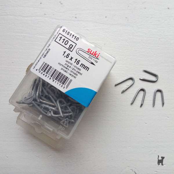 Auf dem Foto liegen vier metallische Krampen auf einer weißen Oberfläche. Daneben liegt ihre Verpackung.