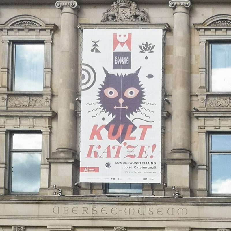 Das Foto zeigt die Fassade des Überseemuseums Bremen. Prominent platziert ein riesiges Banner, dass die Sonderausstellung 'Kult Katze!' bewirbt