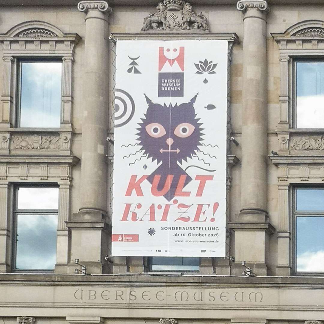 Das Foto zeigt die Fassade des Überseemuseums Bremen. Prominent platziert ein riesiges Banner, dass die Sonderausstellung 'Kult Katze!' bewirbt